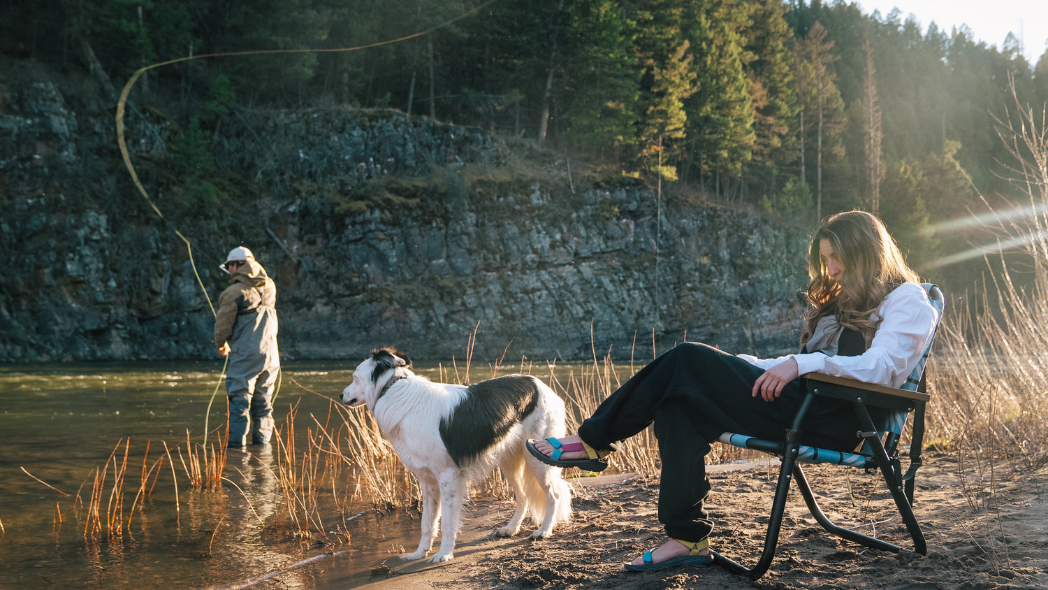 Lifestyle image of a woman relaxing in a camp chair beside a river with a dog nearby, while a man fishes in the water surrounded by forest and rocky cliffs. Woman and man wearing Bickford USA's Overall bibs, Anorak Sailcloth Jacket, and Everyday Long Tee