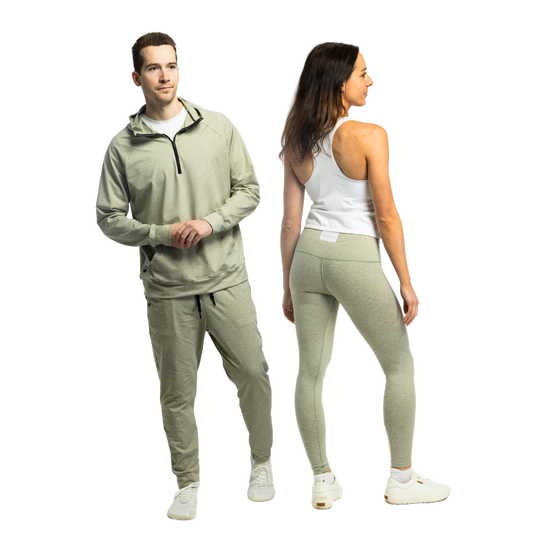 A man and woman standing side by side in matching heather sage green Bickford USA athletic wear — the man wears the Athletic Hooded Quarter Zip with joggers and white sneakers, and the woman is shown from the back in high-waisted leggings paired with a white racerback tank top and white sneakers, against a white background.