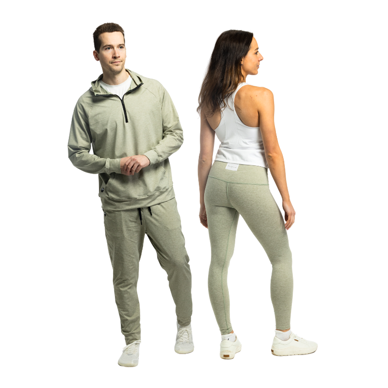 A man and woman standing side by side in matching heather sage green Bickford USA athletic wear — the man wears the Athletic Hooded Quarter Zip with joggers and white sneakers, and the woman is shown from the back in high-waisted leggings paired with a white racerback tank top and white sneakers, against a white background.