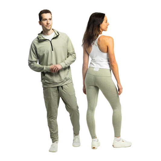 A man and woman standing side by side in matching heather sage green Bickford USA athletic wear — the man wears the Athletic Hooded Quarter Zip with joggers and white sneakers, and the woman is shown from the back in high-waisted leggings paired with a white racerback tank top and white sneakers, against a white background.