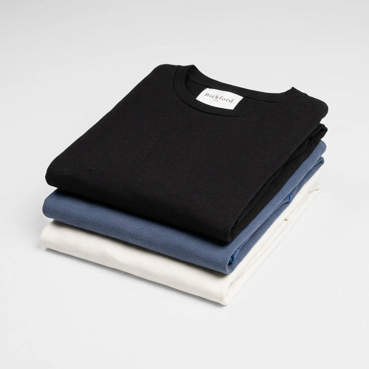 A stack of three folded Bickford USA Everyday Long Sleeve tees — black on top with the cream Bickford USA woven neck label visible, slate blue in the middle, and cream on the bottom — arranged in a neat flat lay against a soft white background.