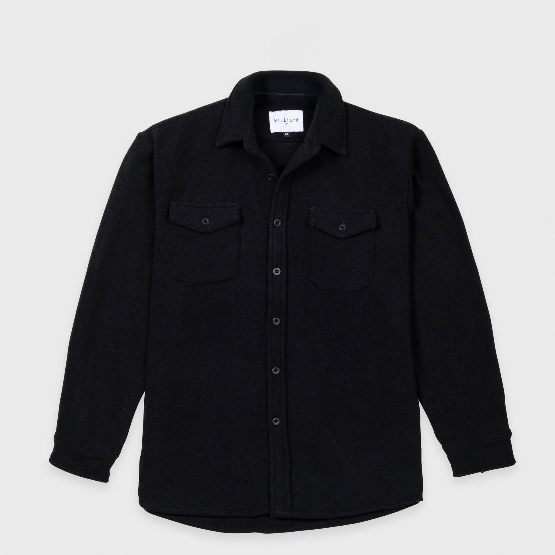 Bickford USA button-down fleece overshirt in black, flat lay front view, featuring a spread collar, dual chest pockets with button flaps, a full front button placket with matte black buttons, and a curved hem, against a light gray background.