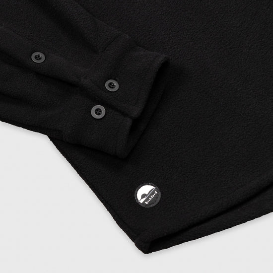 Close-up detail of the cuff and lower hem of the Bickford USA black button-down fleece overshirt, showing the two-button barrel cuff, matte black buttons, and a circular woven Bickford logo patch on the lower left hem.