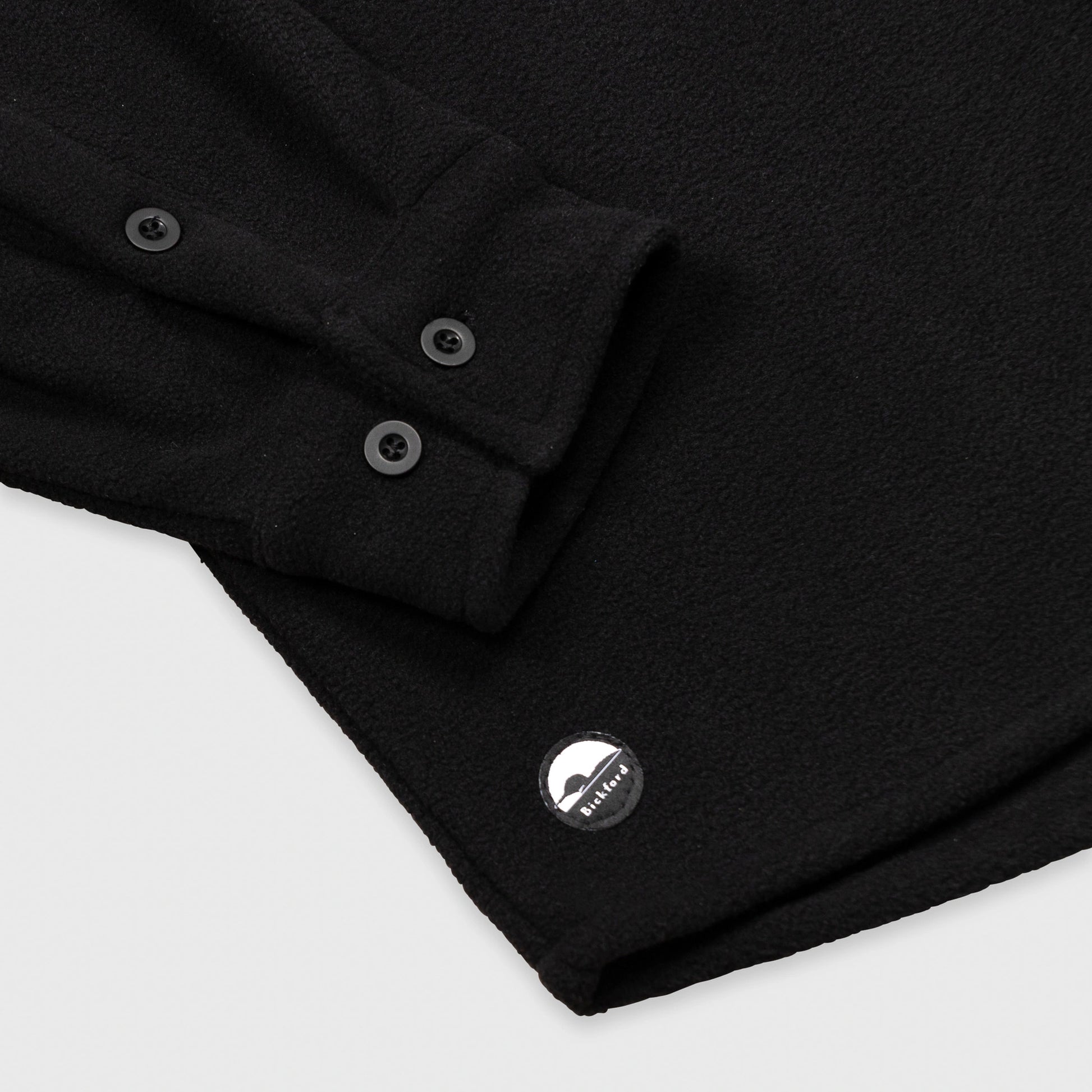 Close-up detail of the cuff and lower hem of the Bickford USA black button-down fleece overshirt, showing the two-button barrel cuff, matte black buttons, and a circular woven Bickford logo patch on the lower left hem.