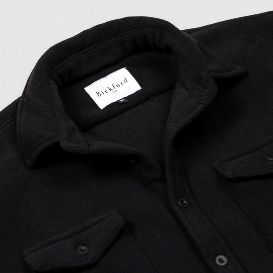 Close-up detail of the open collar and upper chest of the Bickford USA black button-down fleece overshirt, showing the spread collar, Bickford USA size M label inside the neck, matte black buttons, and chest pocket flap.