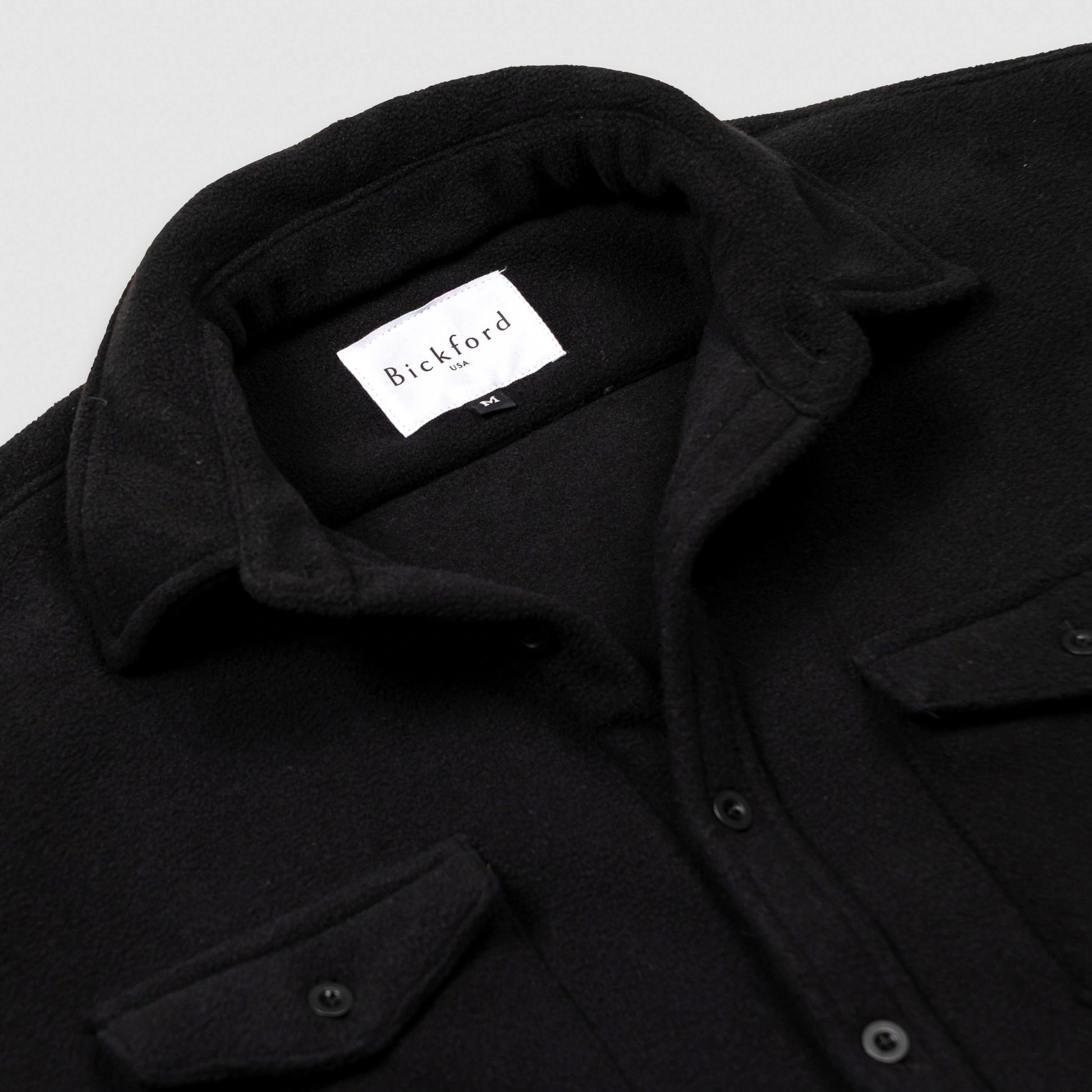 Close-up detail of the open collar and upper chest of the Bickford USA black button-down fleece overshirt, showing the spread collar, Bickford USA size M label inside the neck, matte black buttons, and chest pocket flap.