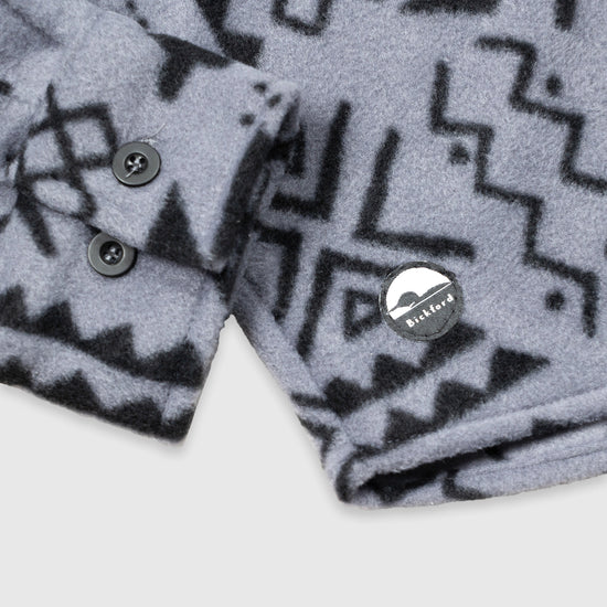 Close-up detail of the cuff and lower hem of the Bickford USA Boulder colorway button-down fleece overshirt, showing the two-button barrel cuff, dark buttons, and a circular woven Bickford logo patch on the lower left hem.