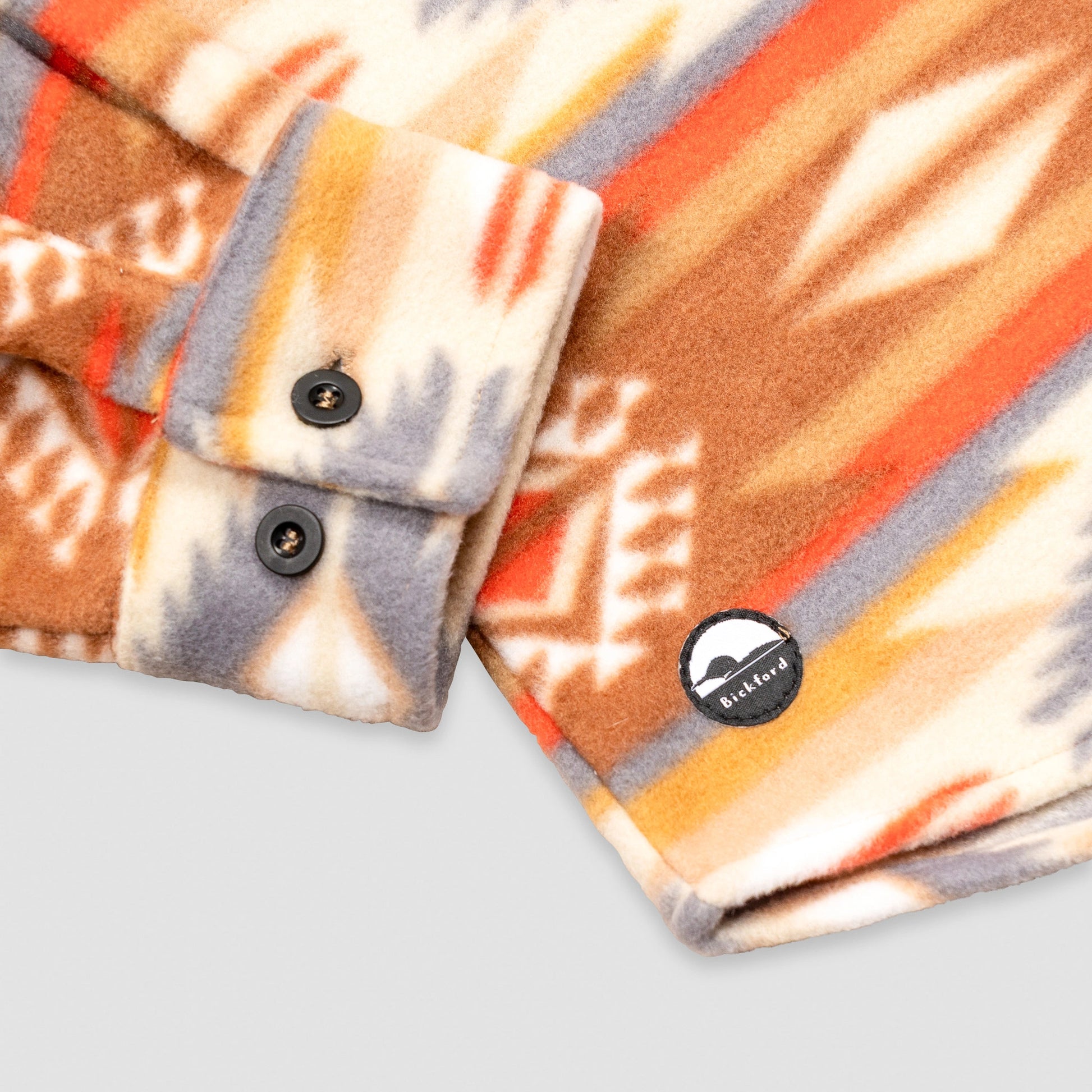 Close-up detail of the cuff and lower hem of the Bickford USA Heritage Trail colorway button-down fleece overshirt, showing the two-button barrel cuff, dark buttons, and a circular woven Bickford logo patch on the lower left hem.