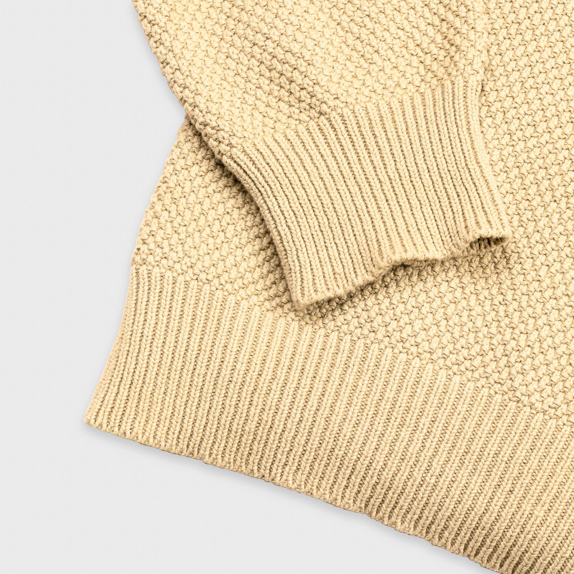 Extreme macro close-up of the cream Fisherman Crewneck fabric, showing the chunky seed stitch knit texture and natural yarn fibers in detail.
