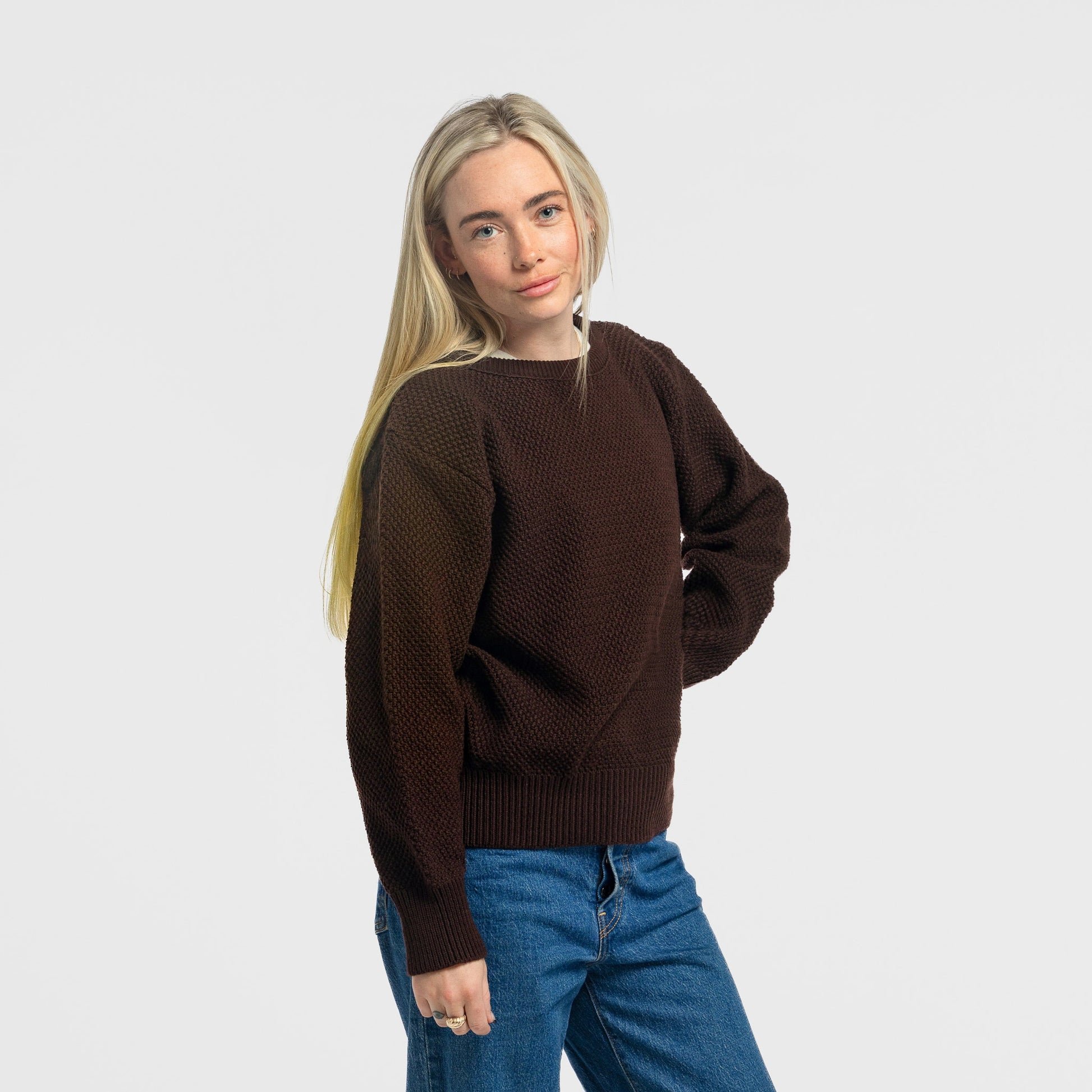 Woman wearing the Bickford USA Fisherman sweater in dark brown, paired with blue straight-leg jeans, shown in a three-quarter pose against a light gray background.