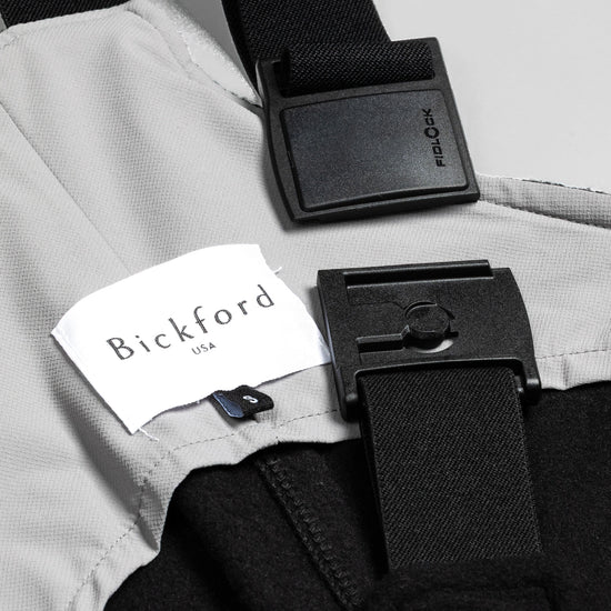 Close-up detail of a Bickford USA label and Fidlock magnetic buckle hardware on the black fleece overall bib, showing the two-piece snap closure system.