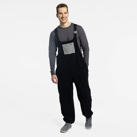 Man wearing the Bickford USA black fleece overall bib over a gray long-sleeve shirt, with gray sneakers, smiling and standing against a white background.