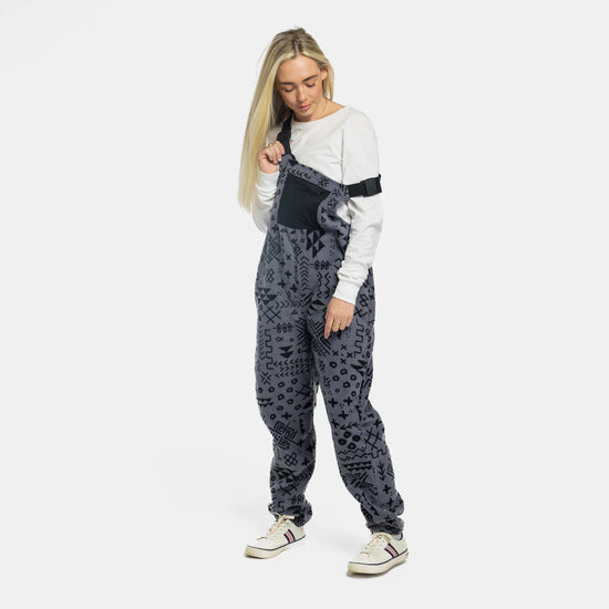 Female model wearing the Bickford USA Boulder colorway fleece overall bib over a white long-sleeve top and white sneakers, adjusting the left shoulder strap, against a white background.