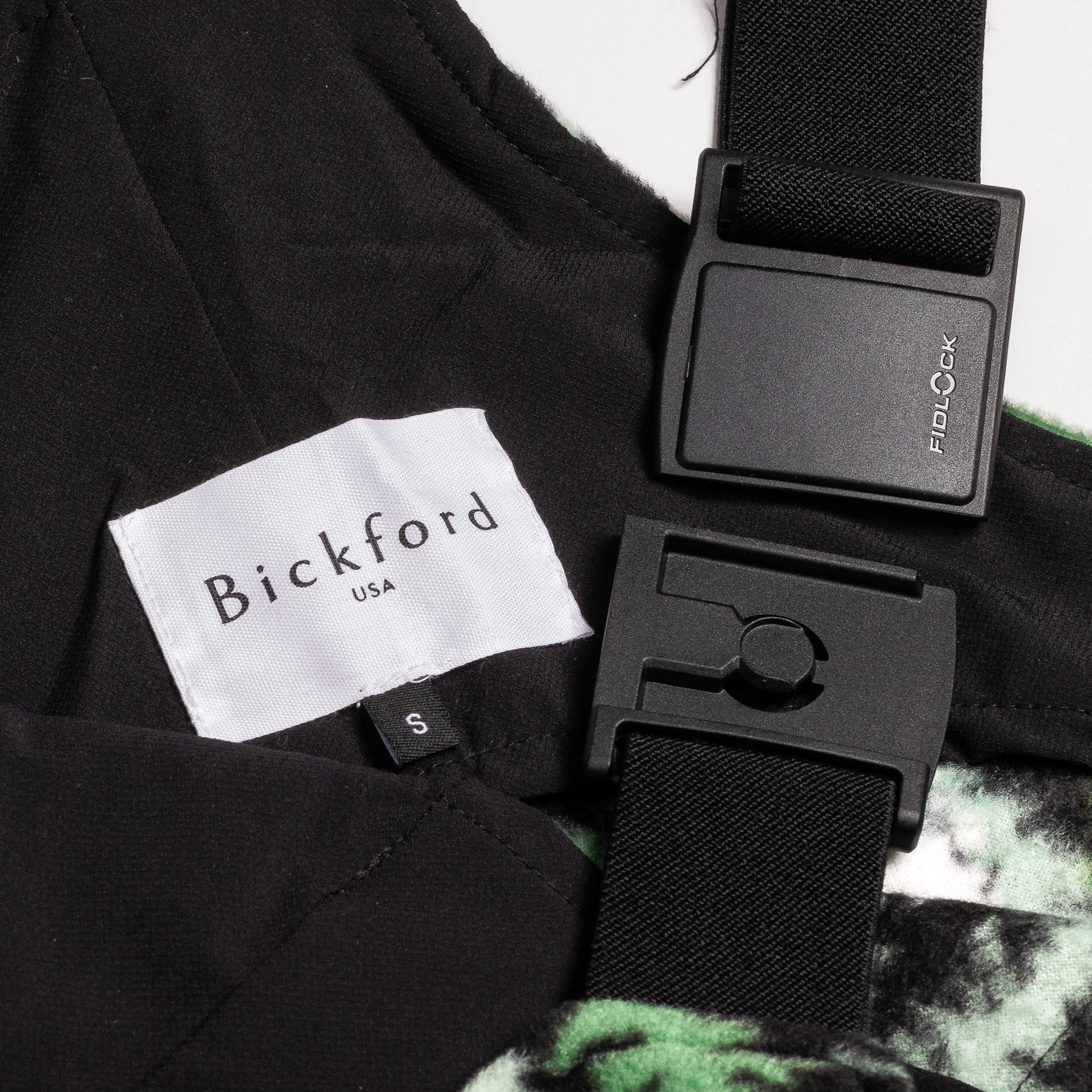 Close-up detail of a Bickford USA label with size S tag and Fidlock magnetic buckle hardware on the Eastern Pine colorway overall bib, with black interior lining and green-print fleece visible.