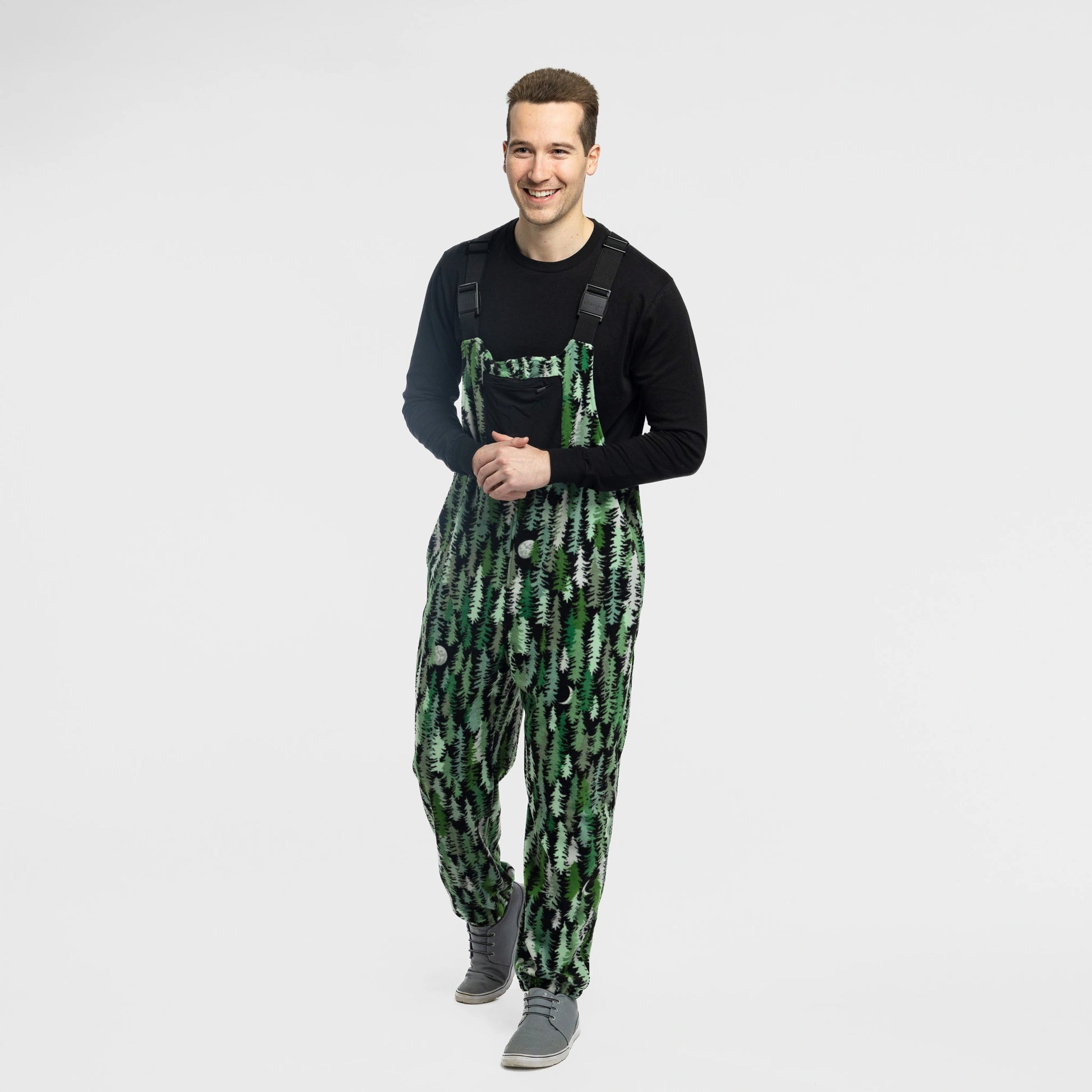 Man wearing the Bickford USA Eastern Pine colorway fleece overall bib over a black long-sleeve shirt, with gray sneakers, hands clasped and smiling, against a white background.