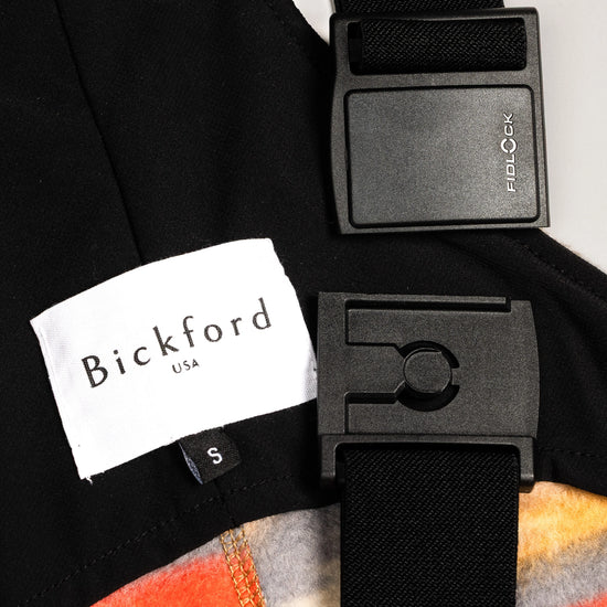 Close-up detail of a Bickford USA label with size S tag and Fidlock magnetic buckle hardware on the Heritage Trail colorway overall bib, with black interior lining and the warm-toned Southwestern print fleece visible.