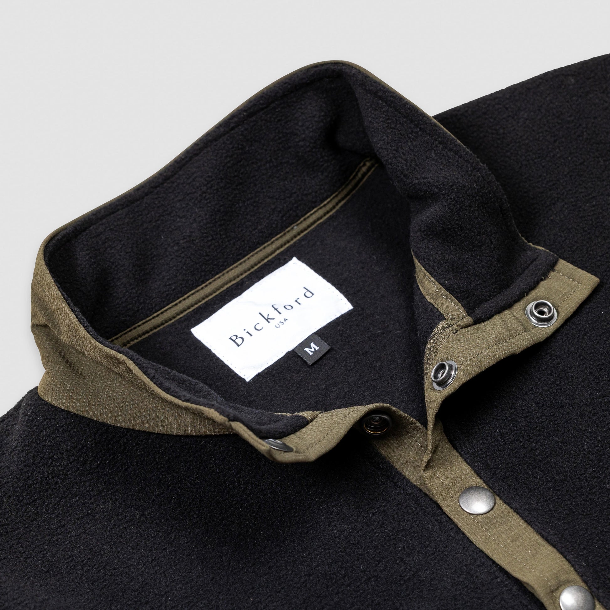 Close-up detail of the collar and snap placket on the Bickford USA black quarter-snap fleece pullover, showing the olive grosgrain trim, silver snap buttons, and Bickford USA size M label inside the collar.