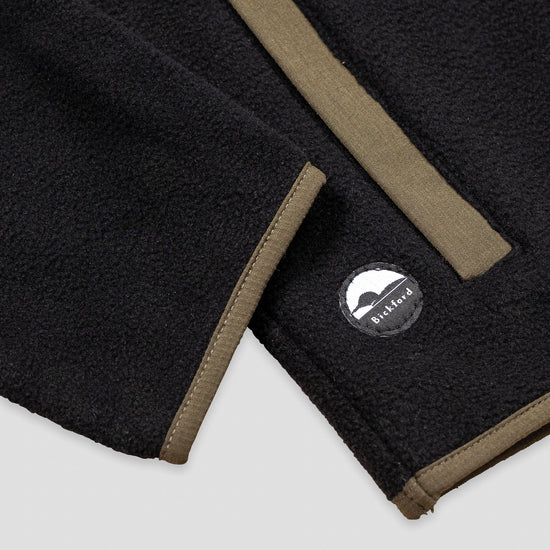 Close-up detail of the hem and side pocket of the Bickford USA black quarter-snap fleece pullover, showing the olive grosgrain pocket trim and a circular woven Bickford logo patch on the lower left hem.