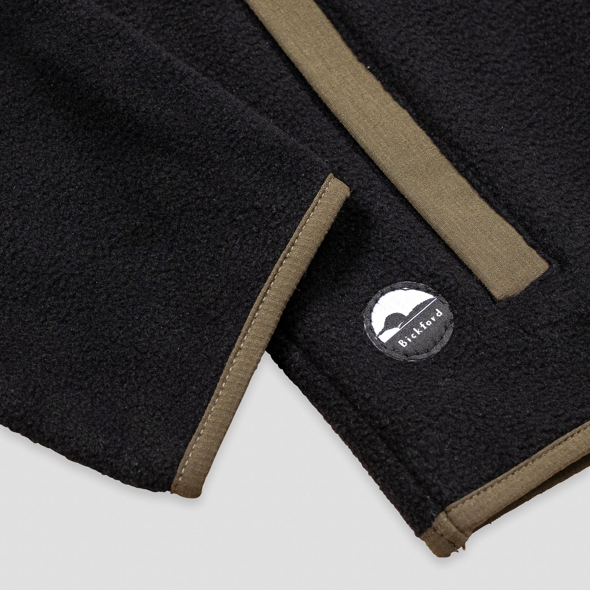 Close-up detail of the hem and side pocket of the Bickford USA black quarter-snap fleece pullover, showing the olive grosgrain pocket trim and a circular woven Bickford logo patch on the lower left hem.