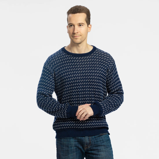 Man wearing Bickford USA The 1926 Sweater in navy and natural, paired with medium-wash denim jeans, shown from the waist up with hands clasped in front against a light gray background.
