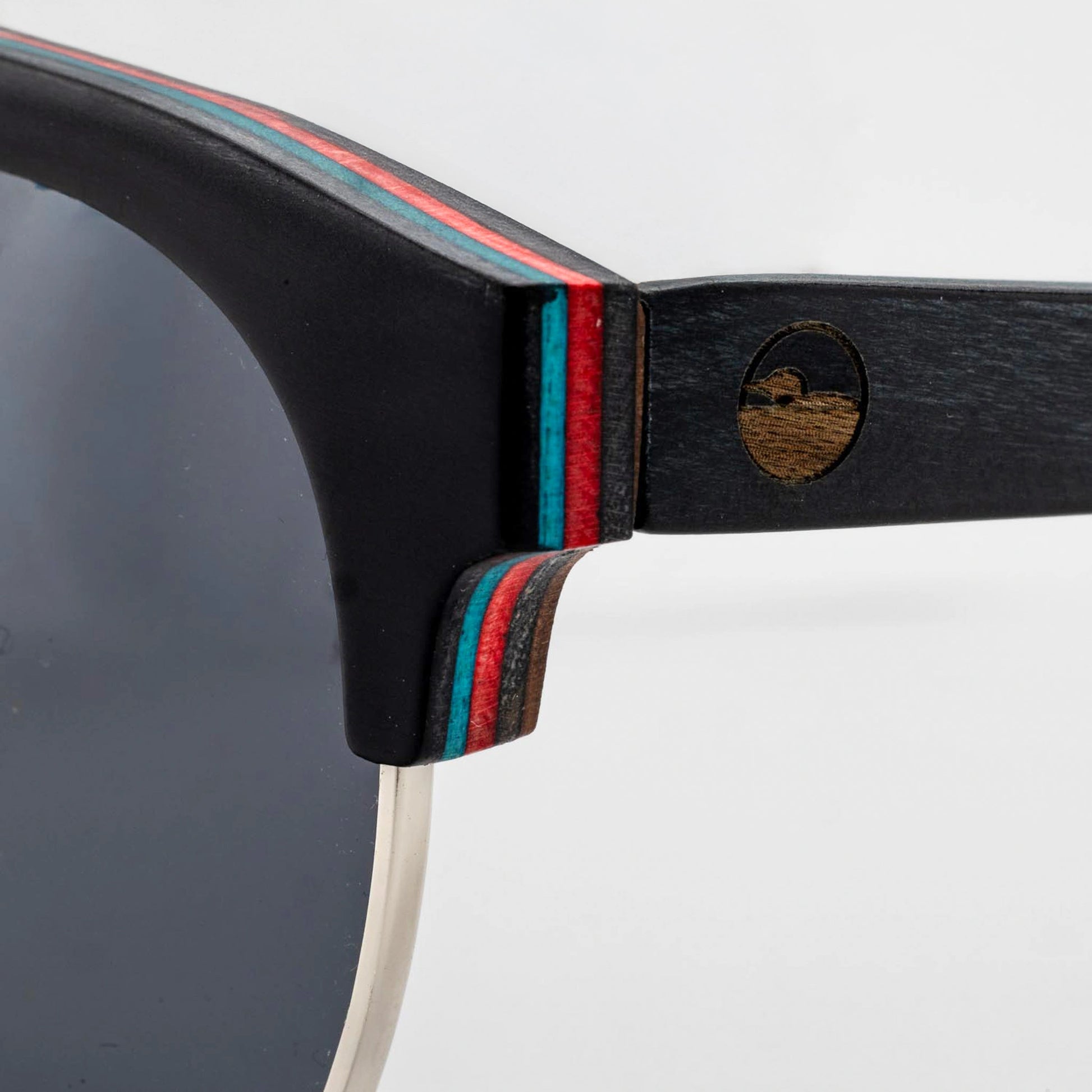 Close-up of the Bickford USA Timberline Half-Frame Sunglasses temple in midnight black, showing the layered red and teal skate-deck wood and engraved loon logo.
