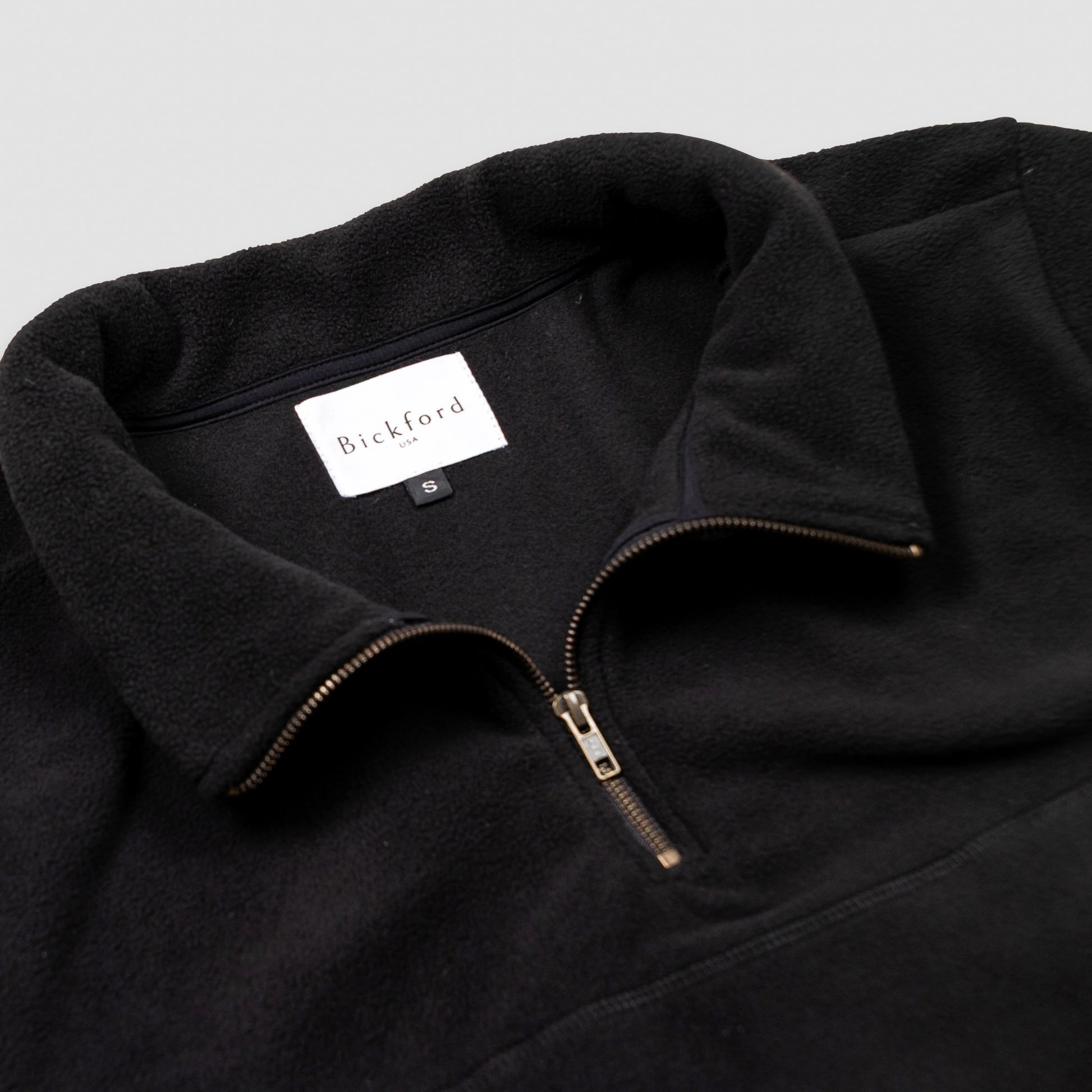Close-up detail of the black Women's Cropped Quarter-Zip showing the open fold-over collar with the Bickford USA label and size tag visible inside, and the brass zipper pull partially unzipped.