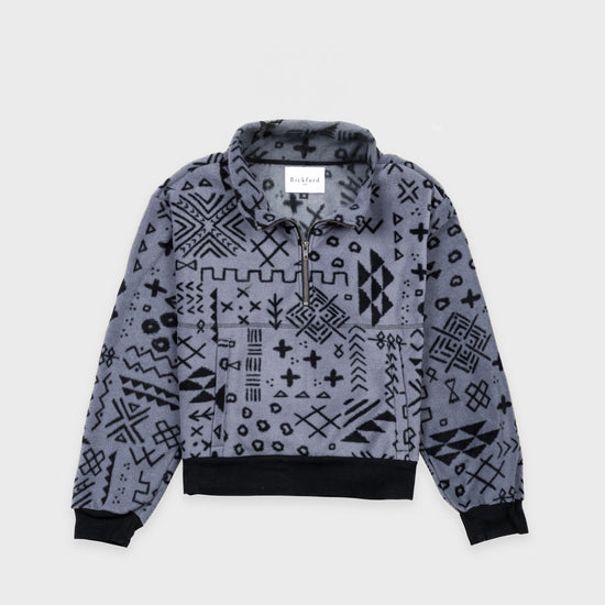 Bickford USA Women's Cropped Quarter-Zip fleece pullover in the Boulder colorway, laid flat on a light gray background, featuring an all-over black geometric tribal print on a grey background, with a wide fold-over collar, brass quarter-zip closure, front kangaroo pocket, and contrasting black ribbed hem and cuffs.