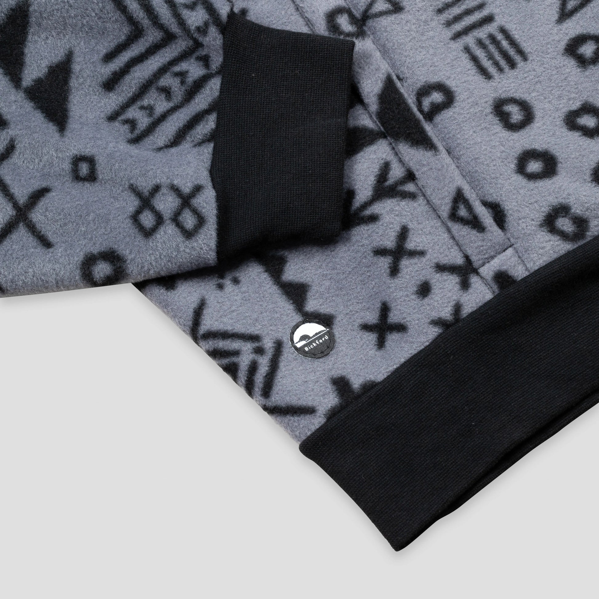 Close-up of the lower hem and kangaroo pocket area of the Boulder Women's Cropped Quarter-Zip, showing the contrasting black ribbed hem band, the pocket opening with a knit tab detail, and the small circular Bickford logo patch against the grey and black geometric print.
