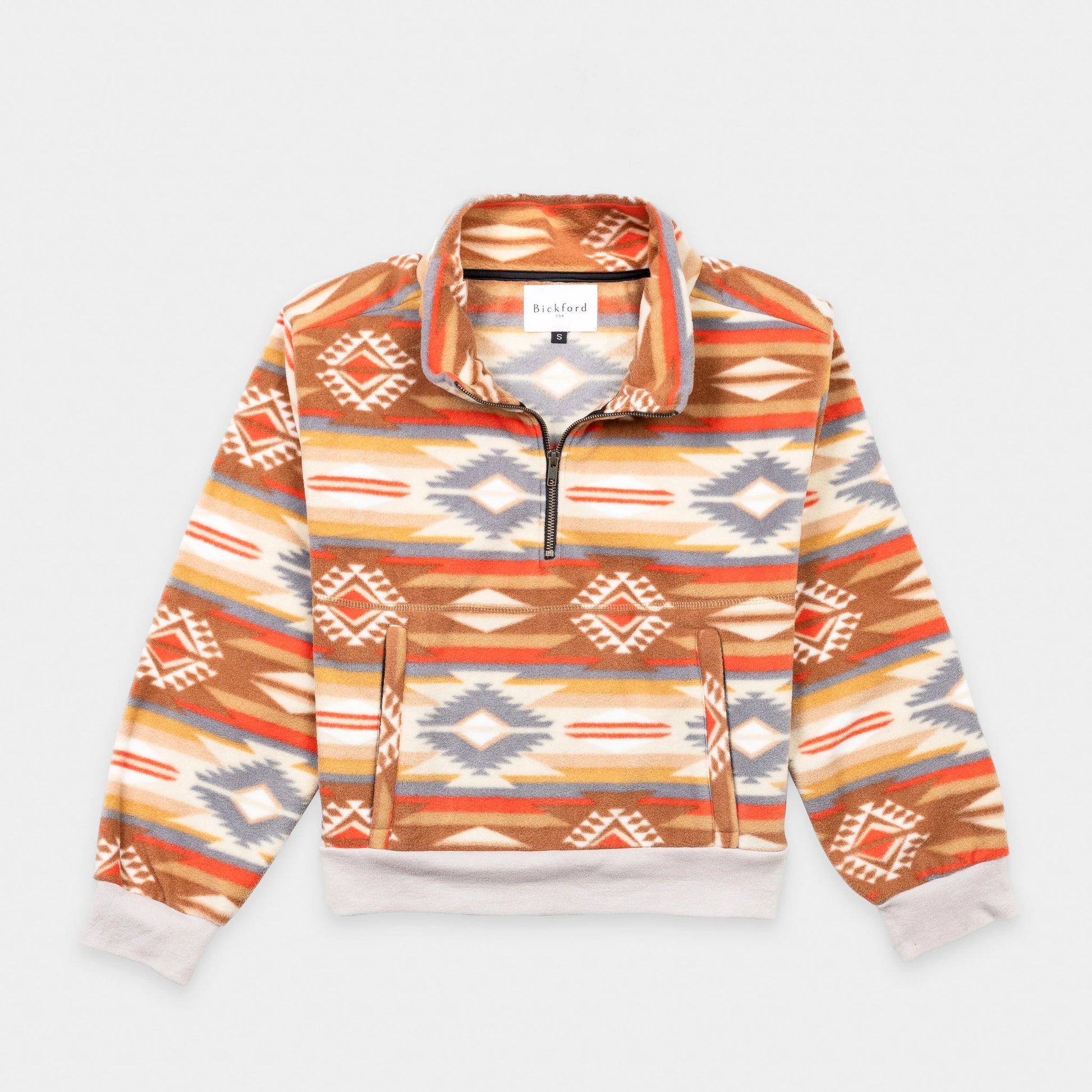 Bickford USA Women's Cropped Quarter-Zip fleece pullover in the Heritage Trail colorway, laid flat on a light gray background, featuring an all-over southwestern-style print with diamond and geometric motifs in orange, brown, red, grey, cream, and gold, with a wide fold-over collar, brass quarter-zip closure, front kangaroo pocket, and contrasting cream ribbed hem and cuffs.