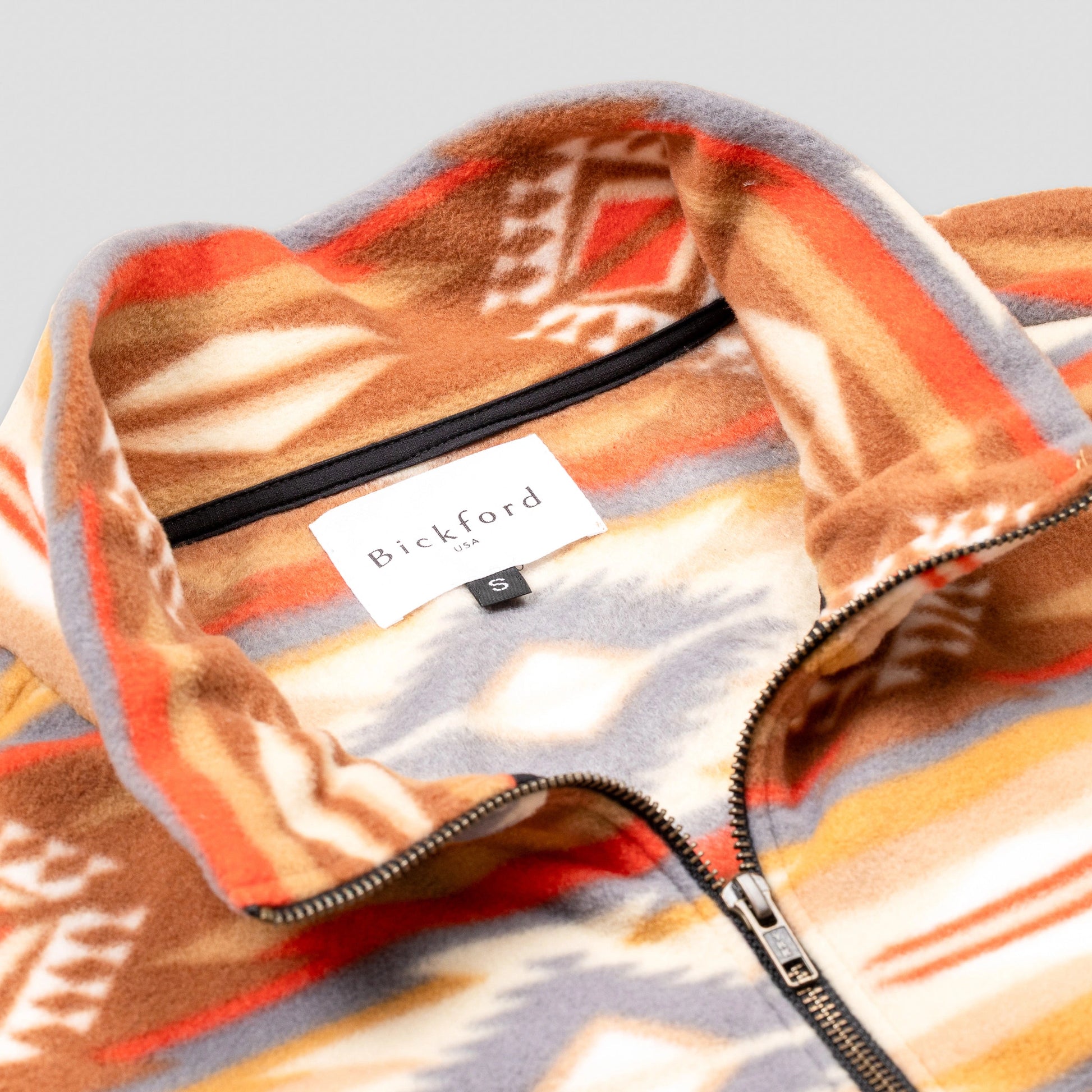 Close-up detail of the Heritage Trail Women's Cropped Quarter-Zip showing the open fold-over collar with the Bickford USA label and size tag visible inside, and the brass zipper pull partially unzipped against the southwestern geometric print in orange, brown, and grey.
