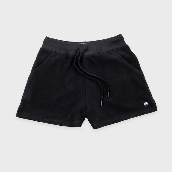 Bickford USA women's fleece shorts in black, flat lay front view, featuring a ribbed elastic waistband, drawstring with barrel-tip ends, side hand pockets, and a circular woven Bickford logo patch on the lower right leg, against a light gray background.