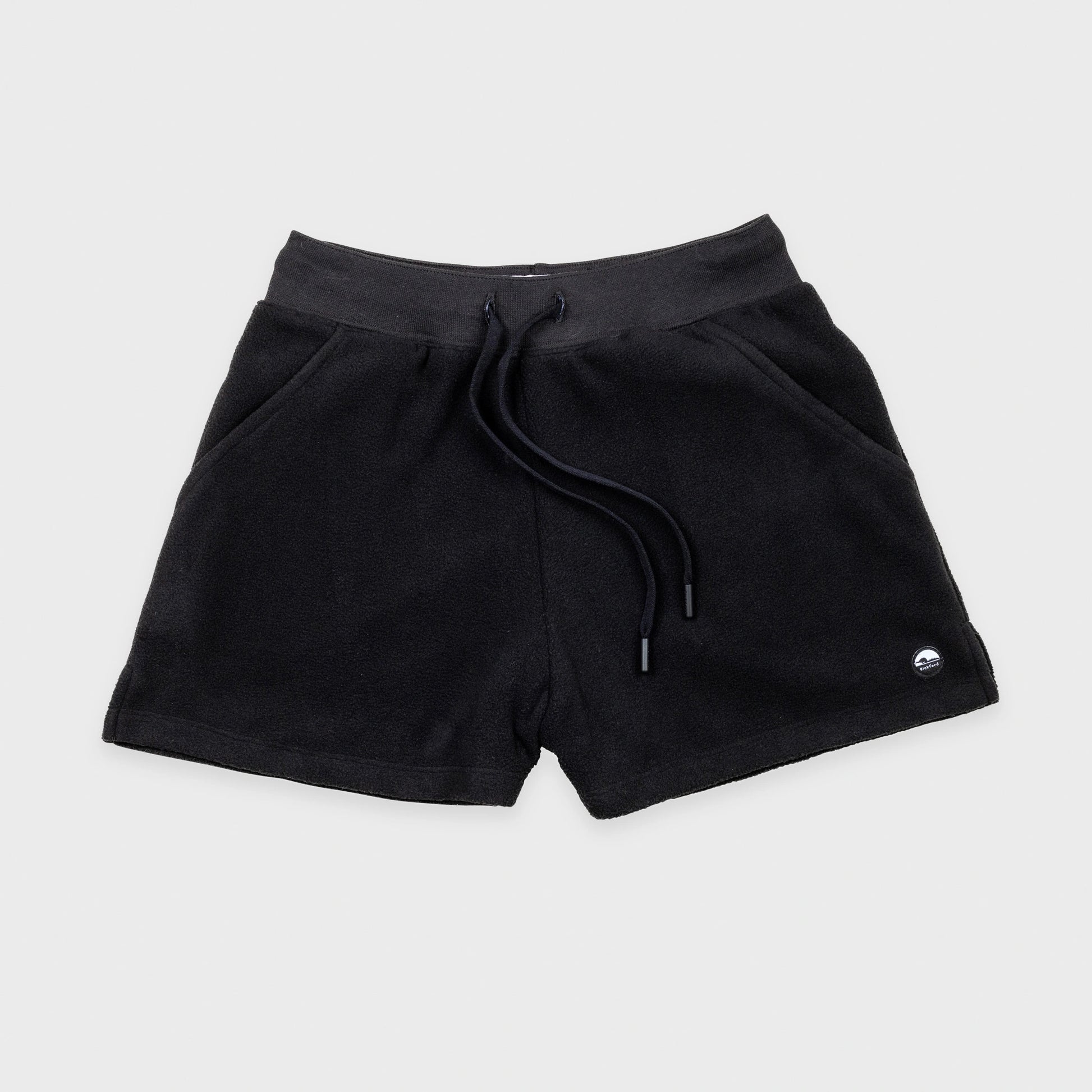 Bickford USA women's fleece shorts in black, flat lay front view, featuring a ribbed elastic waistband, drawstring with barrel-tip ends, side hand pockets, and a circular woven Bickford logo patch on the lower right leg, against a light gray background.