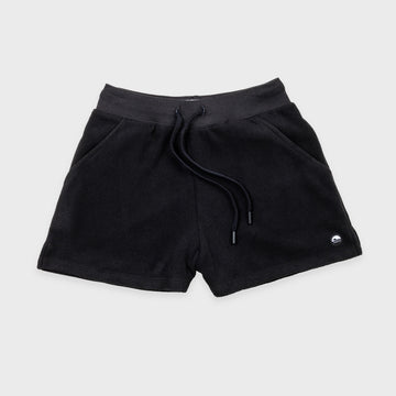 Bickford USA women's fleece shorts in black, flat lay front view, featuring a ribbed elastic waistband, drawstring with barrel-tip ends, side hand pockets, and a circular woven Bickford logo patch on the lower right leg, against a light gray background.