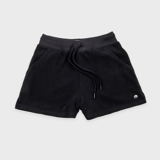 Bickford USA women's fleece shorts in black, flat lay front view, featuring a ribbed elastic waistband, drawstring with barrel-tip ends, side hand pockets, and a circular woven Bickford logo patch on the lower right leg, against a light gray background.