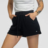 Woman wearing the Bickford USA black women's fleece shorts paired with a white ribbed tank top, hands in pockets, showing the drawstring waistband and side pocket detail, against a white background.