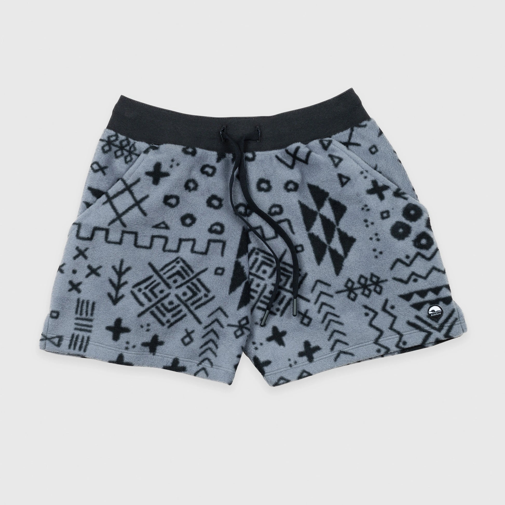 Bickford USA women's fleece shorts in the Boulder colorway — a gray base with an allover black geometric and tribal print — flat lay front view, featuring a black ribbed elastic waistband, drawstring, side hand pockets, and a circular Bickford logo patch on the lower right leg, against a light gray background.