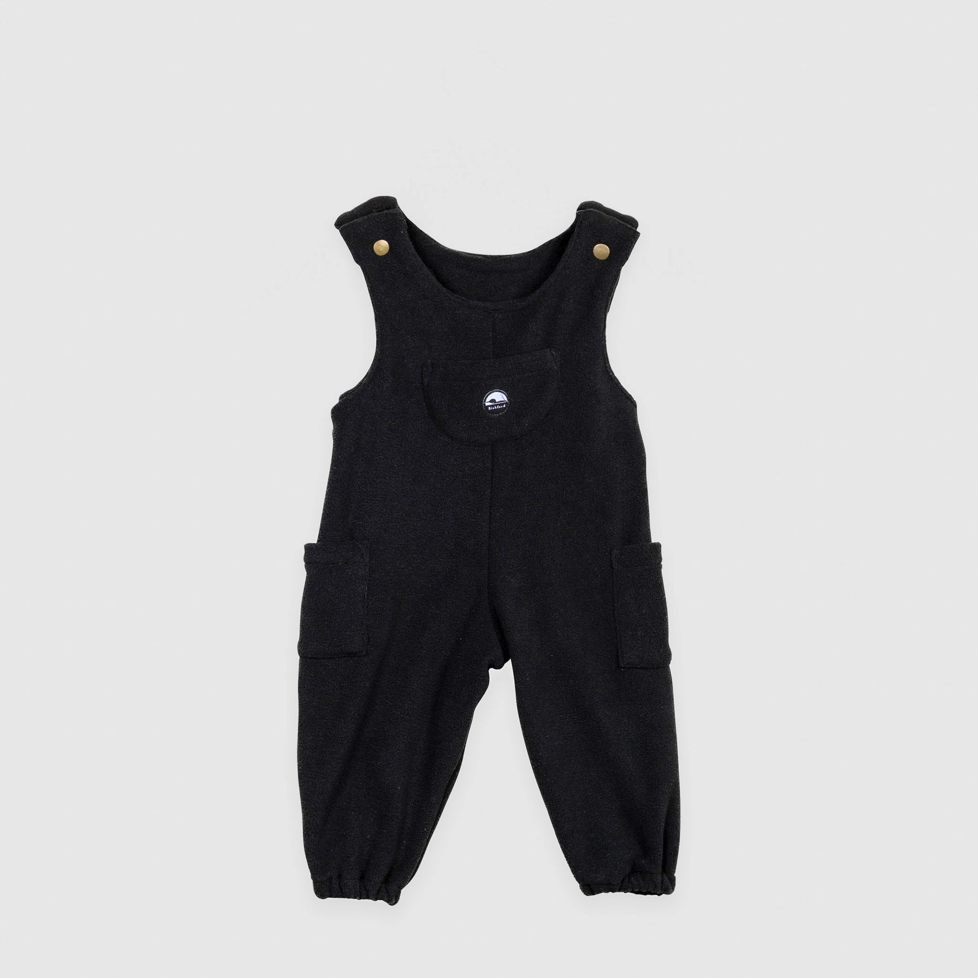 Bickford USA Youth fleece overalls in solid black, laid flat on a light gray background, showing the full front with adjustable shoulder straps with brass snap closures, a chest bib pocket with circular Bickford logo patch, two side cargo pockets, and elasticized ankle cuffs.