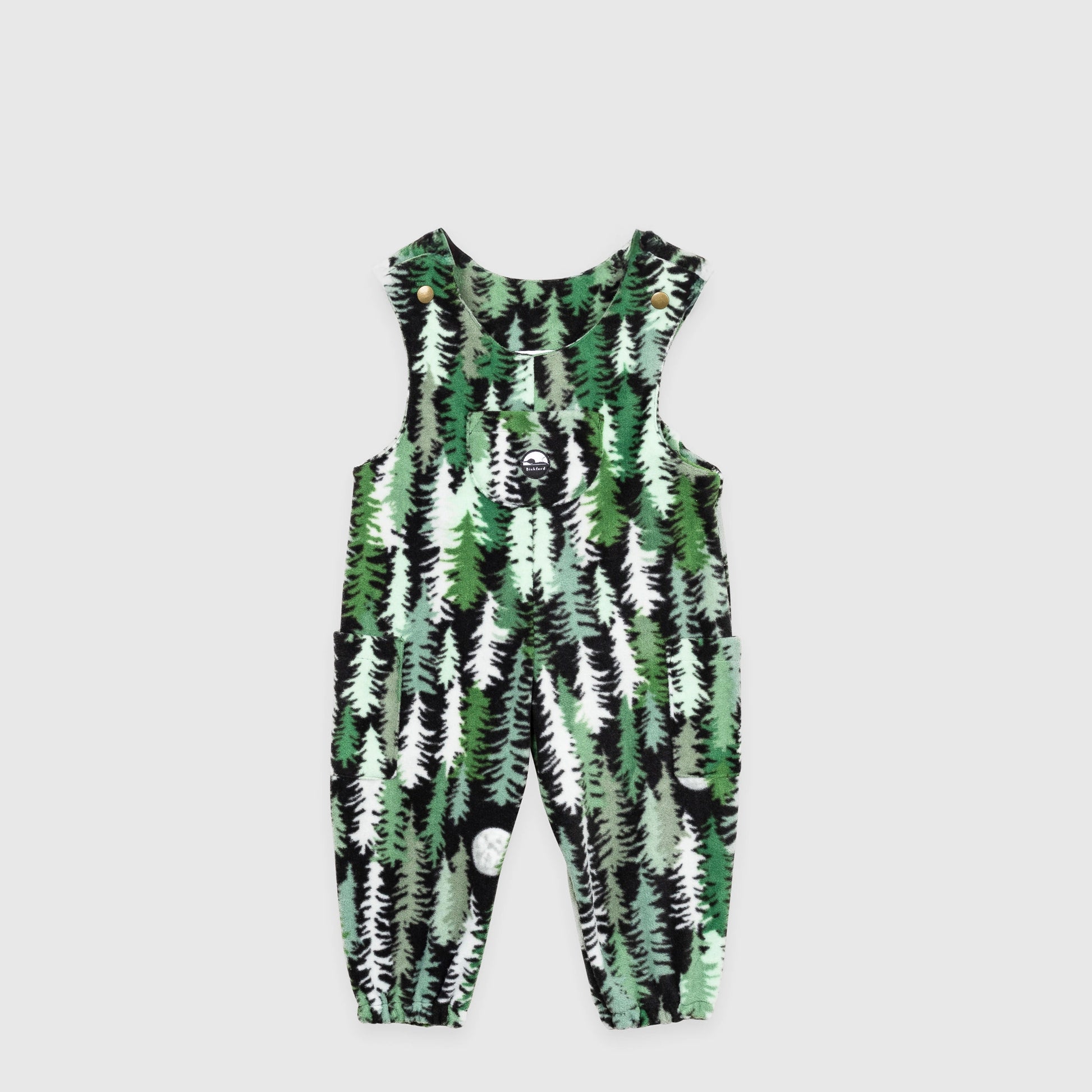 Bickford USA Youth fleece overalls in the Eastern Pine colorway, laid flat on a light gray background, featuring an all-over print of layered pine trees in shades of green, black, and cream with a moon motif, with brass snap shoulder straps, a chest bib pocket with circular Bickford logo patch, two side pockets, and elasticized ankle cuffs.