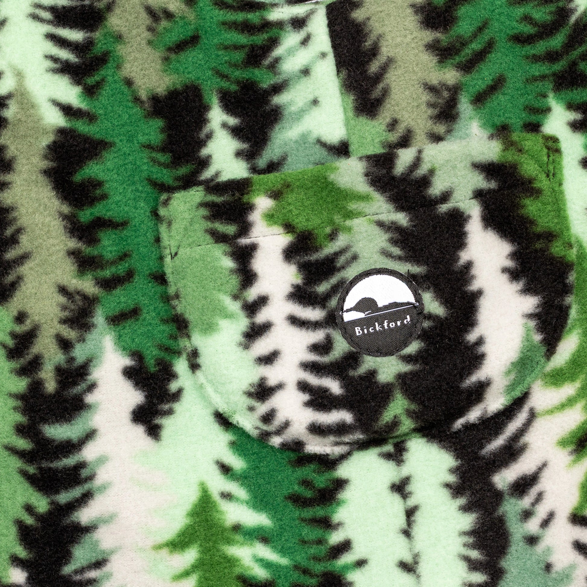 Close-up of the chest bib pocket on the Eastern Pine Youth Overalls showing the circular woven Bickford mountain logo patch centered on the pocket against the pine tree print.