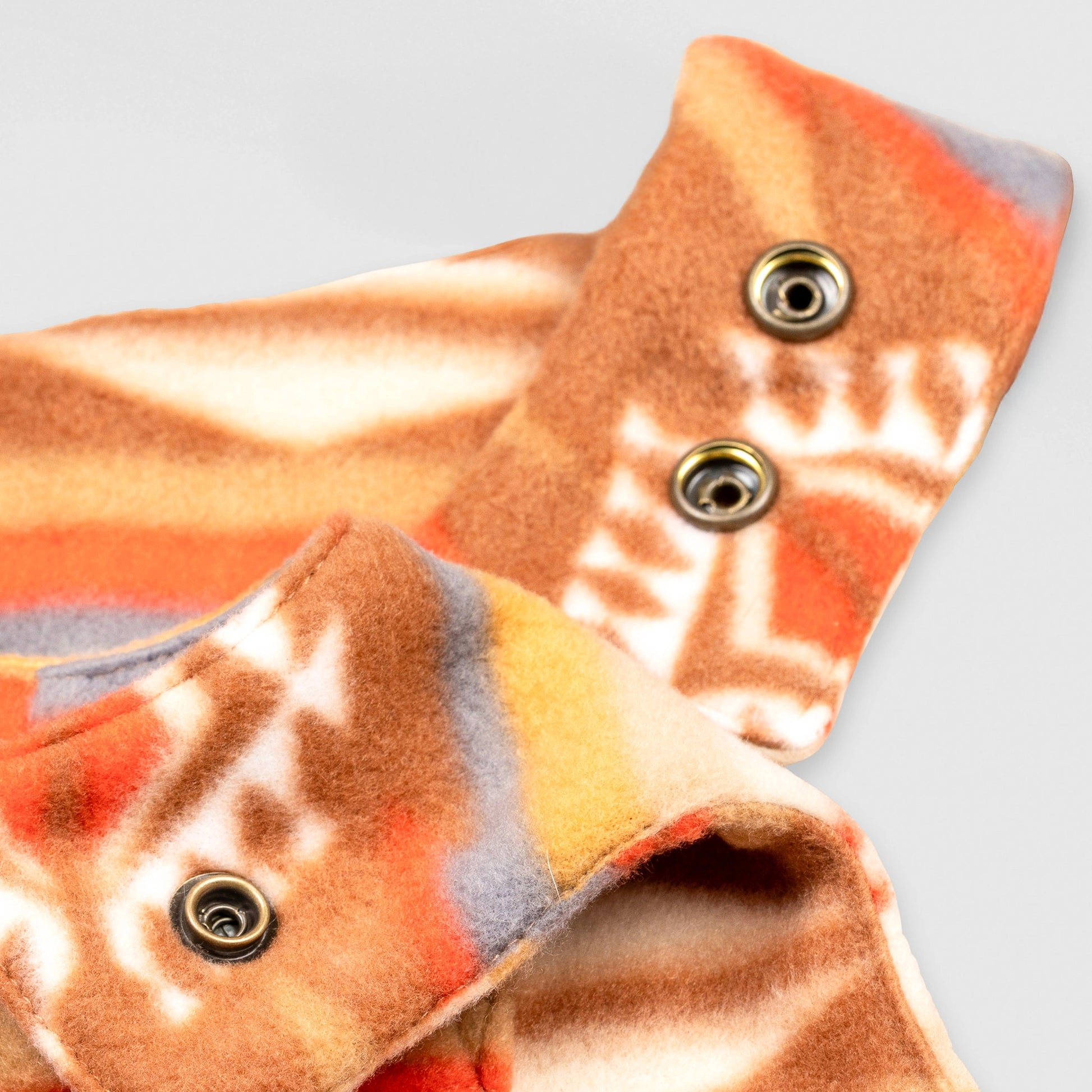 Close-up detail of the Heritage Trail Youth Overalls showing the adjustable shoulder straps with brass snap closures against the southwestern geometric print in orange, brown, and gold.