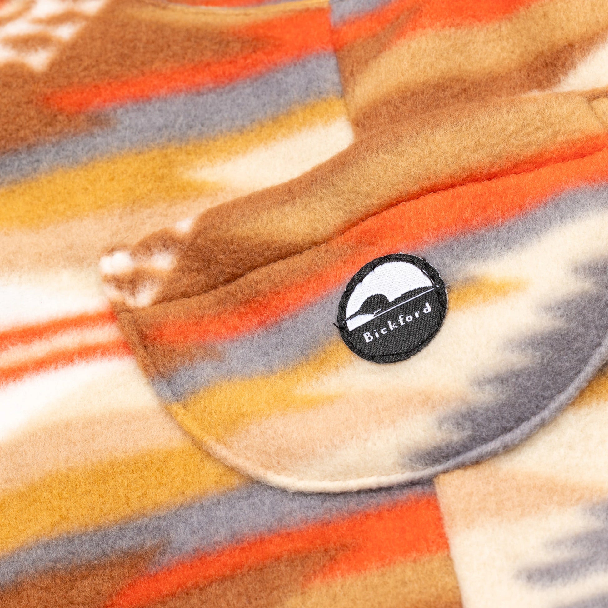 Close-up of the chest bib pocket on the Heritage Trail Youth Overalls showing the circular woven Bickford mountain logo patch against the southwestern stripe and geometric pattern in orange, grey, and gold.