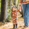 Young toddler walking on a sunlit forest path holding the hand of an adult, wearing the Heritage Trail Youth Overalls in the southwestern geometric print paired with a brown long-sleeve shirt and cowboy boots.