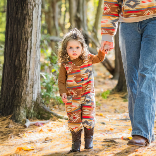 Young toddler walking on a sunlit forest path holding the hand of an adult, wearing the Heritage Trail Youth Overalls in the southwestern geometric print paired with a brown long-sleeve shirt and cowboy boots.