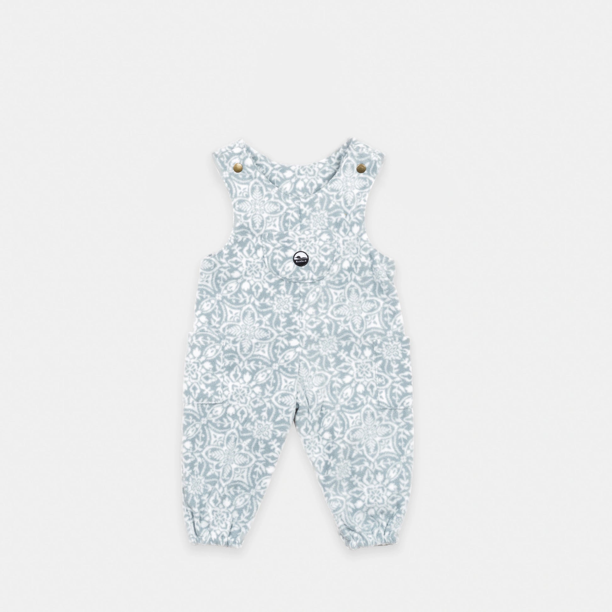 Bickford USA Youth fleece overalls in the Snowflake colorway, laid flat on a light gray background, featuring an all-over ornate snowflake and medallion print in grey and white, with brass snap shoulder straps, a chest bib pocket with circular Bickford logo patch, two side pockets, and elasticized ankle cuffs.