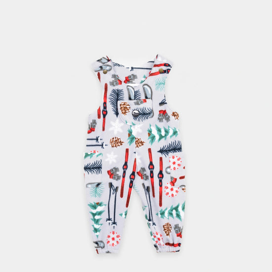 Bickford USA Youth fleece overalls in the Sugarbush colorway, laid flat on a light gray background, featuring an all-over winter sports print with skis, ski poles, ice skates, mittens, snowflakes, pinecones, and pine trees on a light grey background, with brass snap shoulder straps, a chest bib pocket with circular Bickford logo patch, two side pockets, and elasticized ankle cuffs.