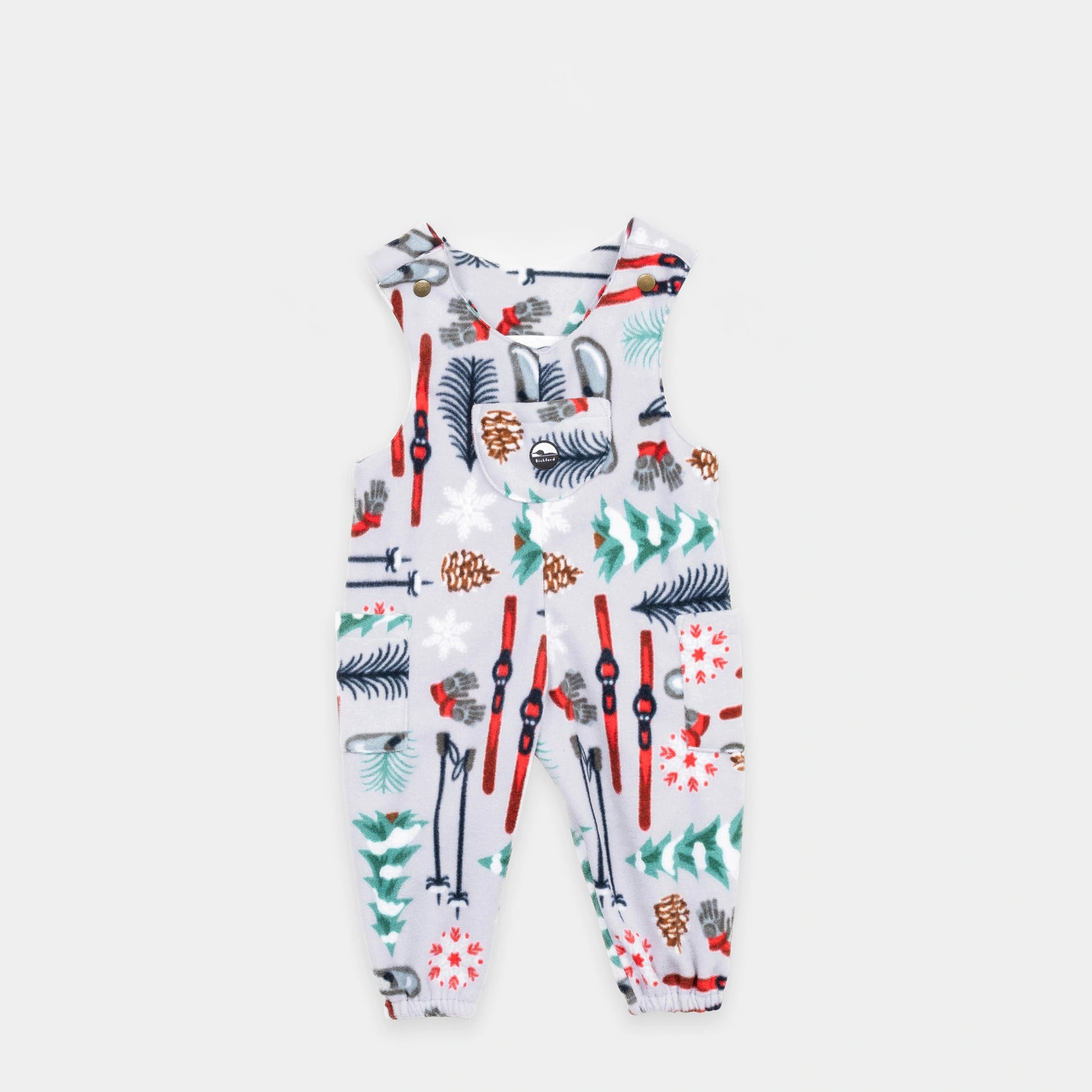 Bickford USA Youth fleece overalls in the Sugarbush colorway, laid flat on a light gray background, featuring an all-over winter sports print with skis, ski poles, ice skates, mittens, snowflakes, pinecones, and pine trees on a light grey background, with brass snap shoulder straps, a chest bib pocket with circular Bickford logo patch, two side pockets, and elasticized ankle cuffs.