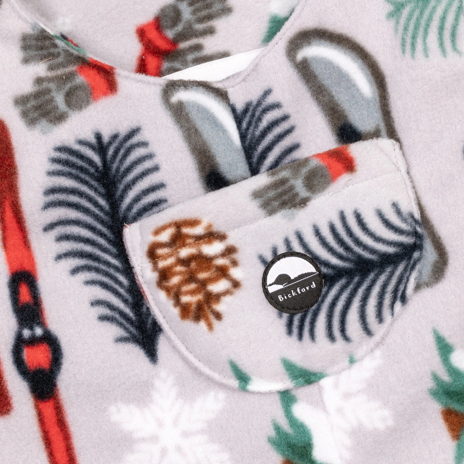 Close-up of the chest bib pocket on the Sugarbush Youth Overalls showing the circular woven Bickford mountain logo patch, with pinecones, pine branches, snowflakes, and skis visible in the surrounding print.