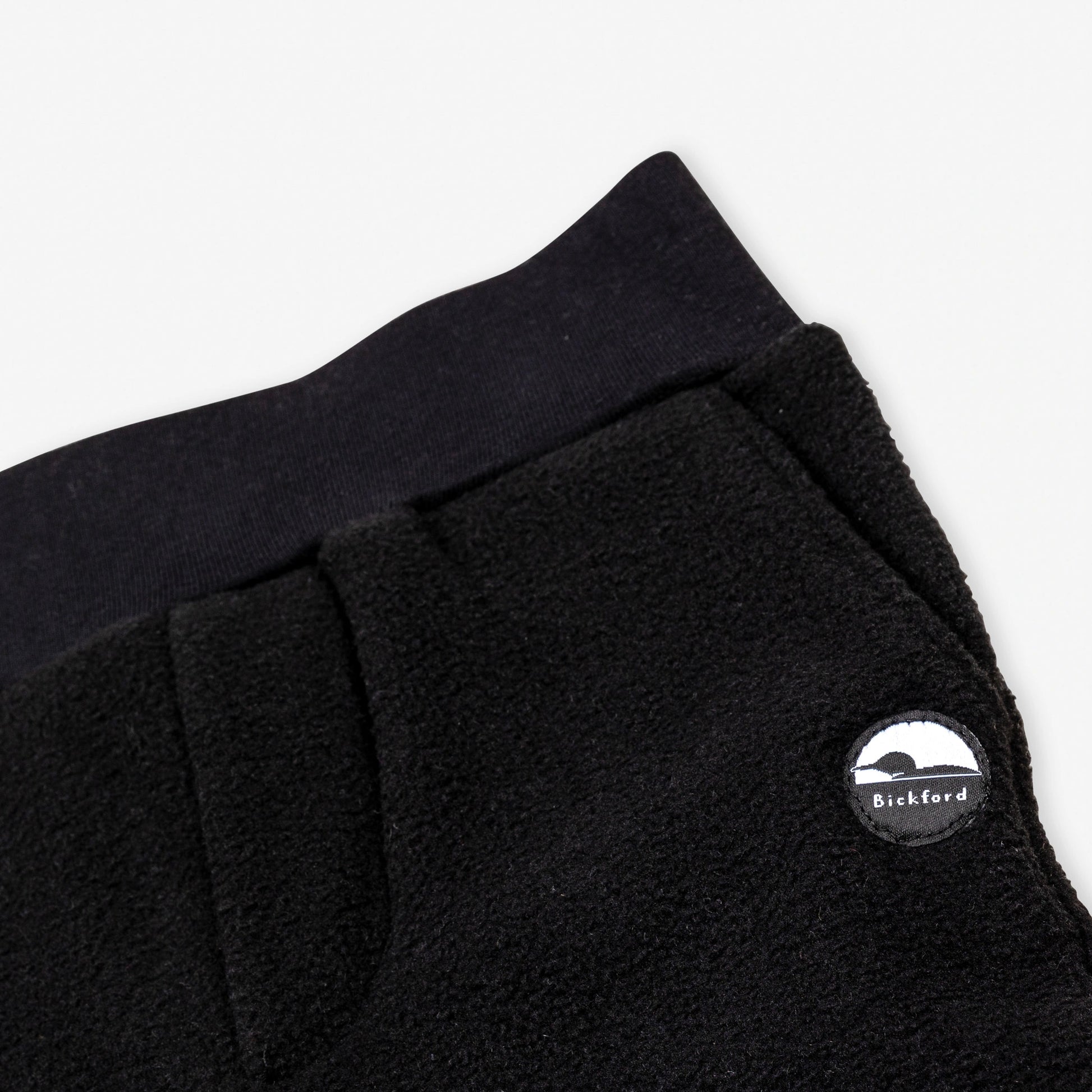 Close-up detail of the black Youth Pants showing the wide black elastic waistband, the angled side pocket opening, and the circular woven Bickford mountain logo patch.


