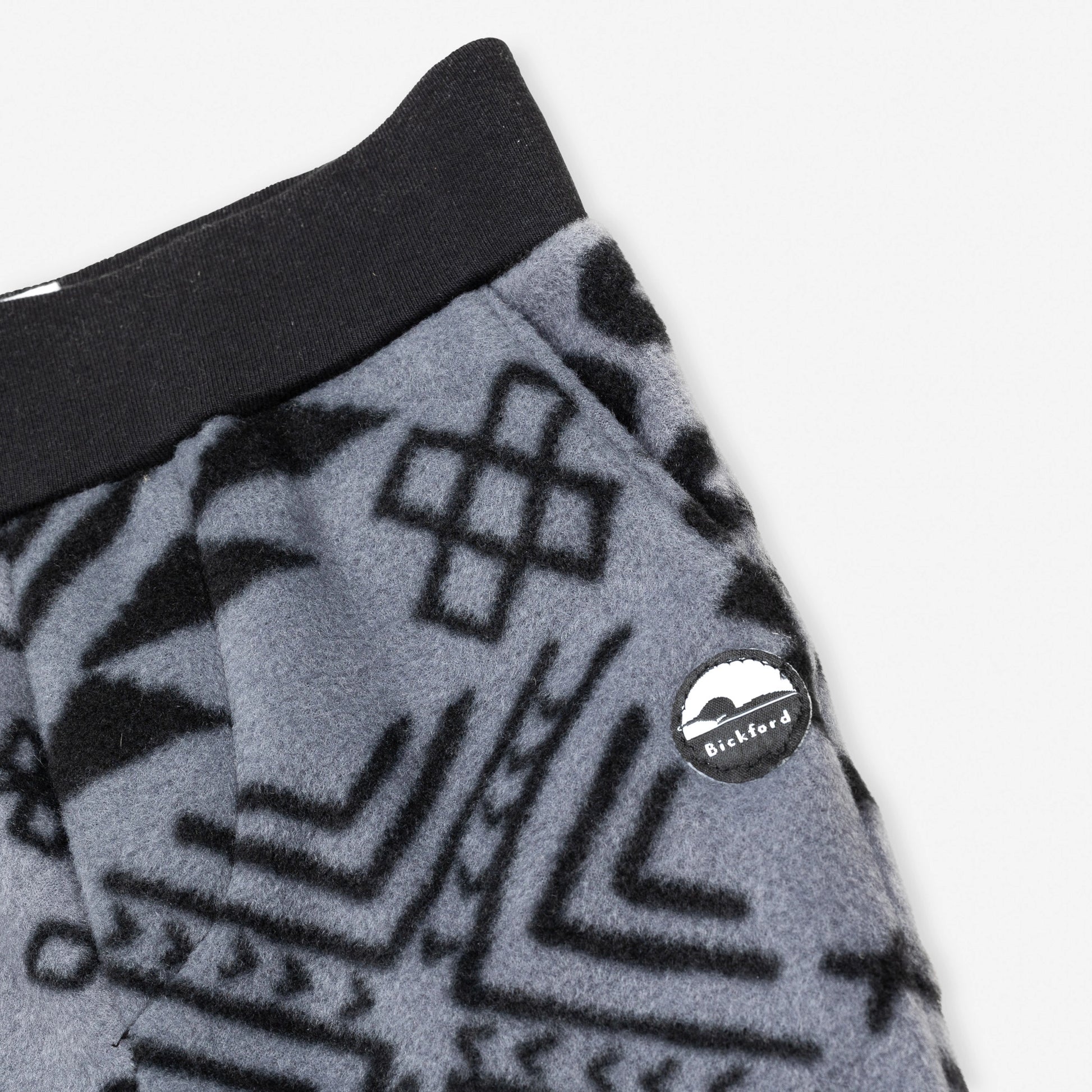 Close-up detail of the Boulder Youth Pants showing the wide black elastic waistband, the angled side pocket, and the circular woven Bickford mountain logo patch against the grey and black geometric tribal print.