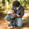 Adult crouching outdoors in dappled sunlight helping a young baby stand upright, with the baby wearing the Boulder Youth Pants in the grey and black geometric tribal print paired with a grey long-sleeve top.